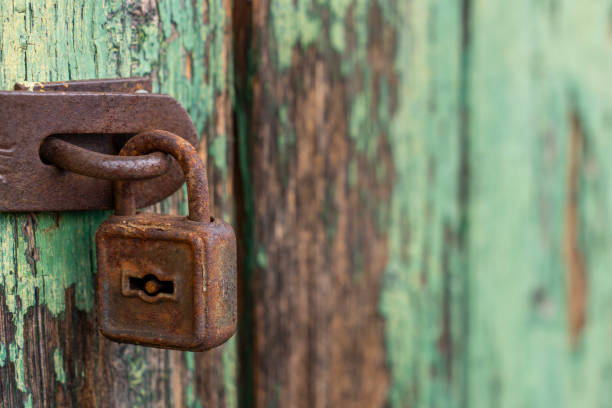 A heavily rusted padlock is attached to a rusty metal hasp on a weathered wooden surface. The wood features peeling green paint, revealing bare patches beneath, adding a rustic, aged look.