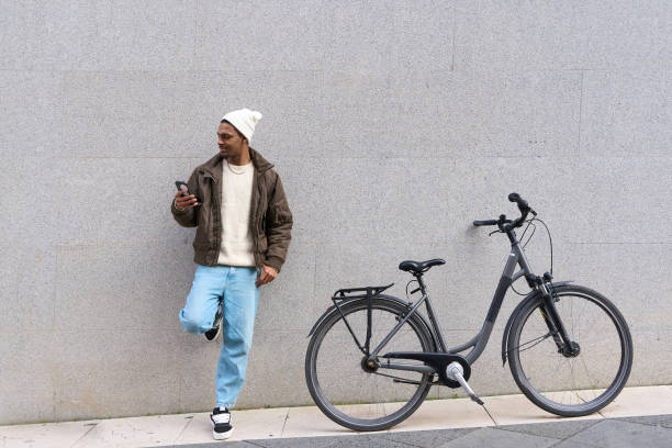 istock Stylish man using smartphone leaning against wall next to his bicycle 2203111209