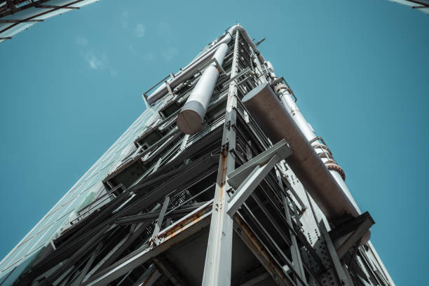 Low-angle view of a modern industrial building with exposed steel framework, pipes, and glass panels. Rusted metal elements contrast with clean lines and a bright blue sky