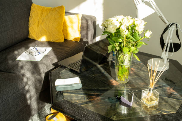 A cozy living room scene with a laptop on a black coffee table, white roses in a glass vase, and yellow pillows on a dark sofa. Sunlight casts soft shadows on the wall.