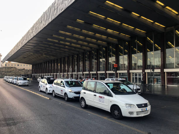A long line of white taxis waiting for passengers in front of Rome Termini. The area is full of coming and going passengers, and taxis are lined up ready to pick up people heading to different parts of the city. The atmosphere is lively, with a constant flow of cars, buses and pedestrians just outside the main entrance to the station.