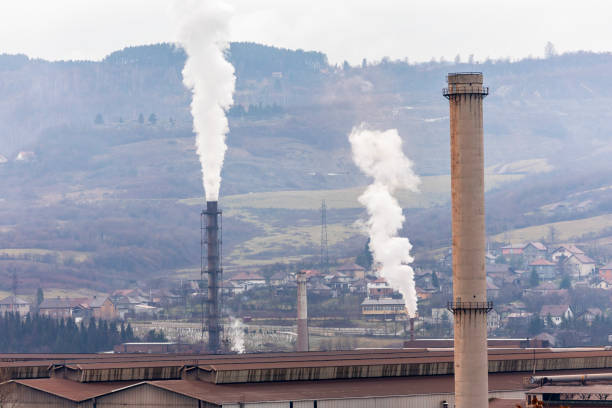 Aerial view of an aging metal factory in Zenica, Bosnia, releasing thick toxic smoke from towering chimneys. Nearby houses stand in the shadow of pollution, highlighting the environmental and health hazards of heavy industry. A powerful visual on air pollution, ecological disaster, and the impact of economic recession on local communities.