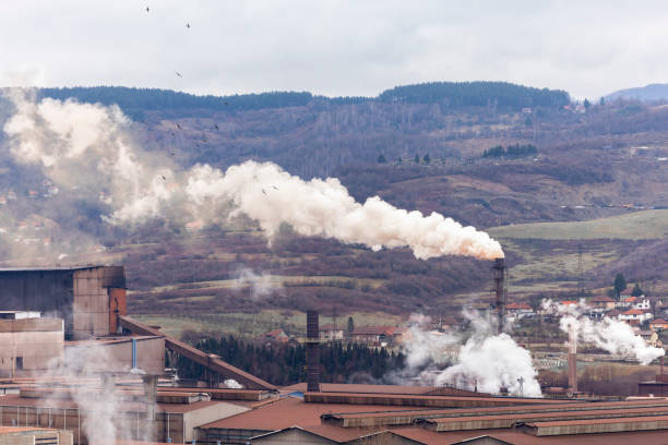 Aerial view of an aging metal factory in Zenica, Bosnia, releasing thick toxic smoke from towering chimneys. Nearby houses stand in the shadow of pollution, highlighting the environmental and health hazards of heavy industry. A powerful visual on air pollution, ecological disaster, and the impact of economic recession on local communities.