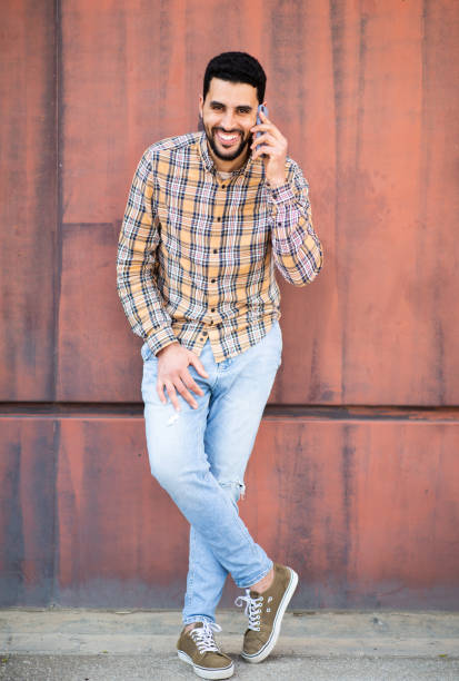 Full length portrait of smiling young arabic man talking on cell phone standing against brown wall