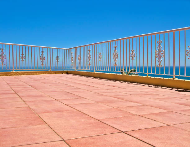 Spacious tiled terrace with pink flooring under a bright blue sky and bordered by a white decorative railing overlooking a tranquil ocean. An ideal setting for relaxation and enjoyment of peaceful coastal sunsets.