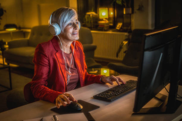 A mature Caucasian woman with short gray hair focuses on her desktop computer in a cozy home office, wearing headphones and a red blazer during late-night work.
