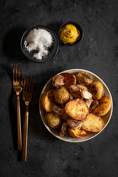 A plate of crispy roasted potatoes with garlic, served with mustard and salt on a dark textured background