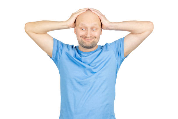 Studio shot of confused bald man holding head with hands, grimacing and looking down