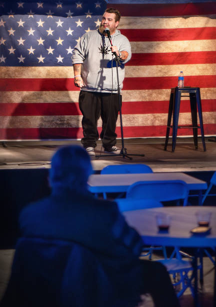 istock Bearded performer with an authentic body type  speaks at event. Middle-aged man delivers speech in front of American flag backdrop with audience seated at blue tables. 2203159694