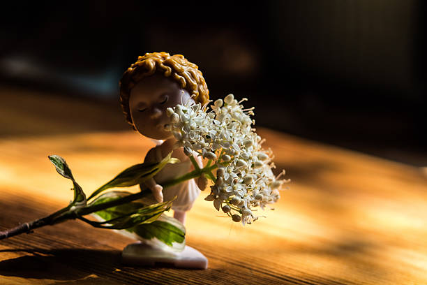 this picture shows a small figure of a baby holding a sprig of flowers. The flowers are white with delicate pink hues and bright anthers. The atmosphere in the photo is cozy and touching, with a soft light that illuminates the figurine and flowers. The scene is reminiscent of spring and the freshness of nature.