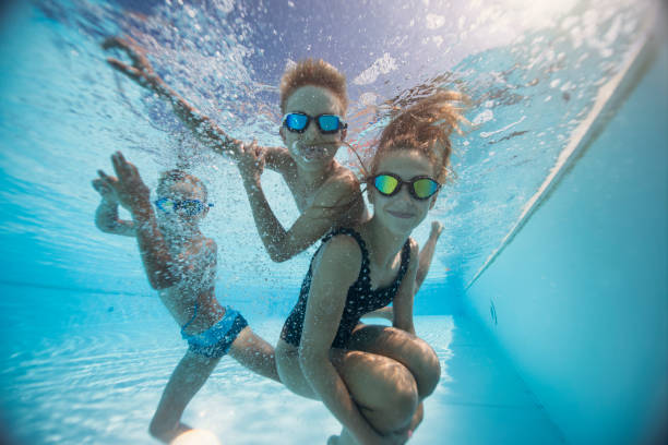 A girl and her two young boys have just jumped into the pool, they are swimming and playing underwater in a clear blue pool. The water is full of bubbles.Sunlight filters through the water, creating shimmering reflections.Shot with Nikkor 14-24 on D850.