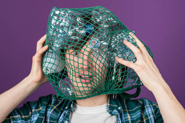 istock Man struggling to remove a green plastic bag filled with empty bottles from his head while expressing concern about plastic pollution 2203193855