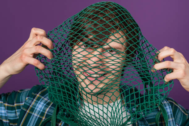 istock Male model sporting plaid shirt, grinning while obscuring face with green mesh net, standing before vivid purple backdrop, conveying whimsical youthful energy 2203194281