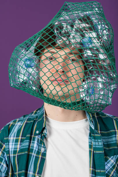 istock Young male wearing plaid shirt standing with plastic bottle net draped over head, symbolizing environmental waste and societal impact of plastic pollution 2203195539
