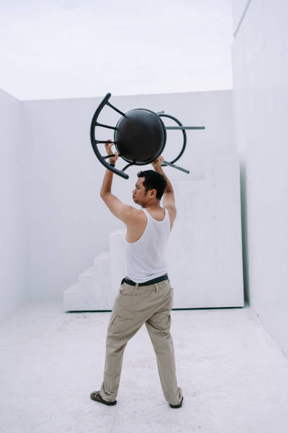 A man lifts a metal chair overhead demonstrating physical strength in a minimalist white architectural space. The scene showcases a blend of physical exertion and modern design, set against a stark, clean backdrop.
