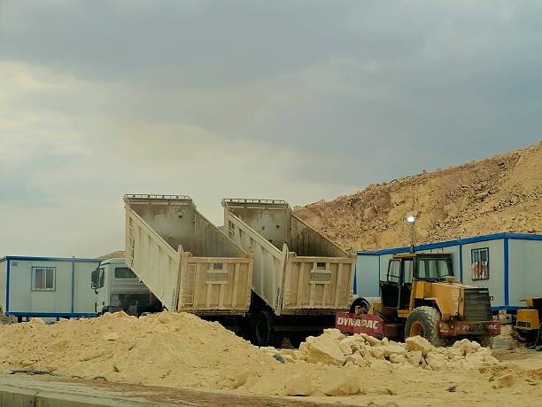 Overhead perspective of a construction site featuring gravel piles and a dump truck loaded with rocks, marked by distinct tire tracks on a sandy terrain