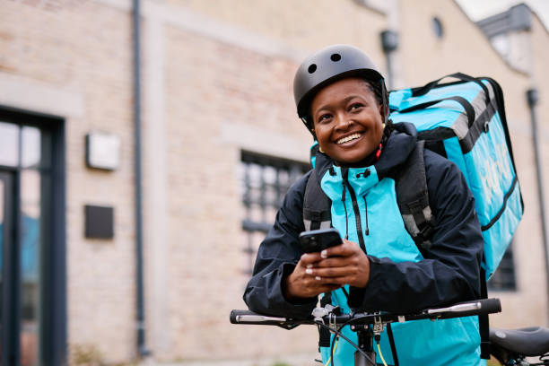 istock Smiling delivery woman using smartphone and carrying backpack on her bicycle 2203197564