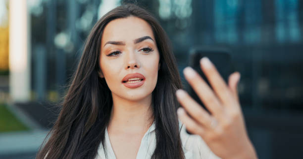 istock A confident woman in a white shirt stands by a railing, chatting through a video call with a friend, the glass office building reflecting the city behind her. 2203199436