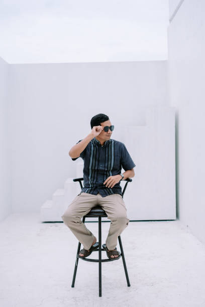 A stylish man wearing sunglasses poses confidently while sitting on a stool against a minimalist white background. His modern attire and confident gaze create a fashionable and eye-catching image, perfect for illustrating contemporary lifestyles and urban aesthetics.