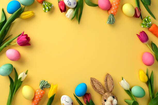 istock Happy Easter concept. Frame of colorful Easter eggs, tulips, and a cute bunny on a yellow background 2203203390