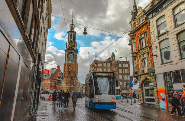 Vibrant cityscape of Amsterdam, Netherlands, captures a classic blue and white tram moving through the bustling streets, framed by historic Dutch architecture. The iconic Munttoren (Mint Tower), with its distinctive clock face and bell tower, stands tall under a dramatic cloudy sky. Pedestrians walk along the wet cobblestone streets, reflecting the lively energy of the city. Overhead tram cables crisscross the scene, emphasizing Amsterdam’s seamless blend of history and modern urban transport