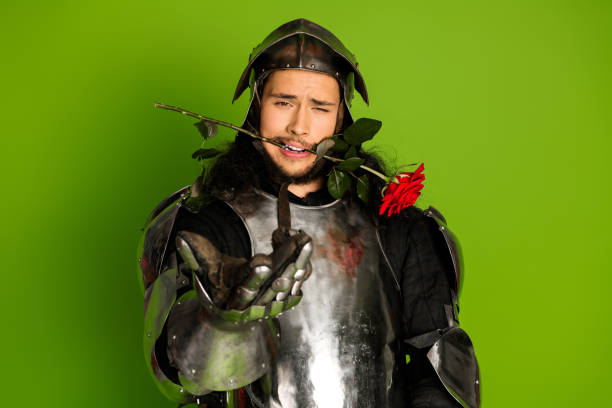 istock Young man in medieval armor biting rose with a playful expression against green background 2203206015