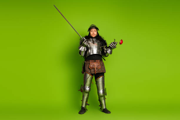istock Medieval knight in armor posing atypically with a rose and sword on a vibrant green backdrop for a whimsical and humorous display 2203206016