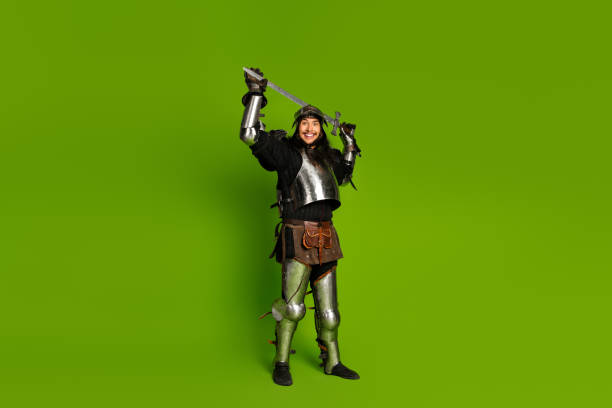 istock Young man in medieval knight armor posing with sword on green background. 2203206089