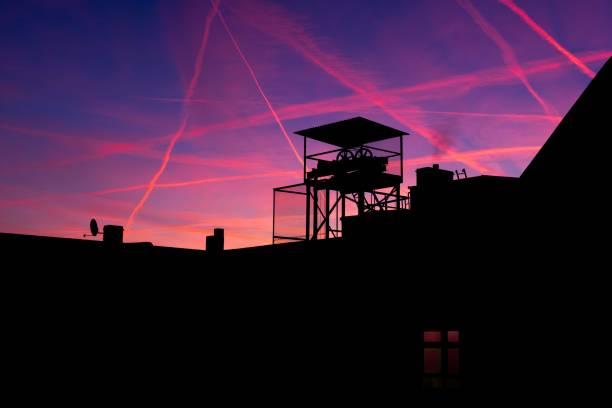 This image captures a sunset over a city skyline, with prominent condensation trails from airplanes crisscrossing the colorful sky. The sky displays a range of warm, saturated colors typical of a sunset, primarily purples and pinks. A central feature is a silhouetted structure, an old elevator platform, with a more intricate design than the surrounding rooftops.