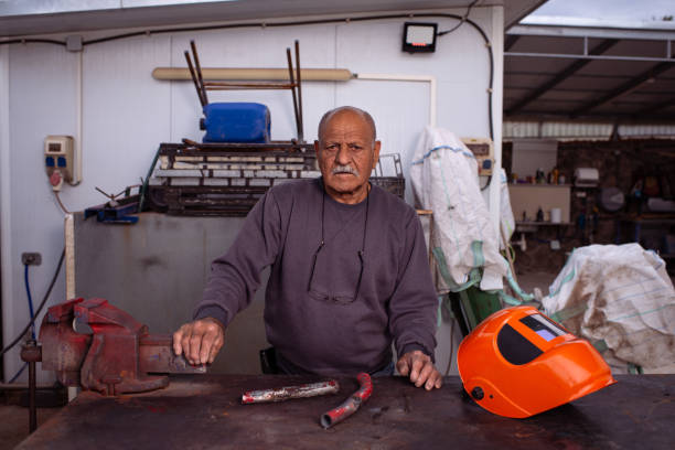 A senior welder with a serious and focused expression stands at his workbench, resting one hand on a metal vise while his protective orange helmet lies beside him. The industrial workshop setting, with tools, pipes, and equipment in the background, emphasizes his years of experience and dedication to metalworking. His worn hands and confident stance reflect a lifetime of craftsmanship and skilled labor.