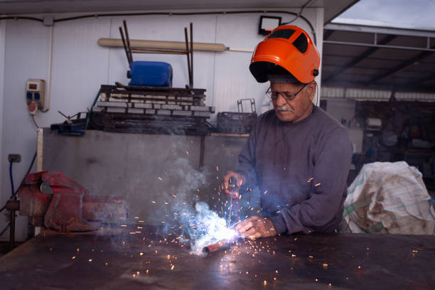 A senior welder, wearing an orange protective helmet raised above his head, carefully welds a metal pipe in an industrial workshop. Sparks fly as he focuses intently on his work, with blue smoke rising from the welding torch. His experienced hands and determined expression reflect a lifetime of dedication to metal craftsmanship. The background, filled with tools and equipment, adds authenticity to the hardworking atmosphere.