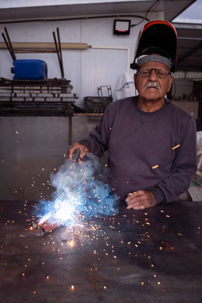 A senior welder, wearing a lifted orange protective helmet and glasses, is skillfully welding metal pipes in a workshop. Sparks fly as blue smoke rises from the intense heat of the welding torch. His steady hands and focused expression highlight years of experience and dedication to craftsmanship. The industrial background, filled with tools and equipment, adds authenticity to the working environment.