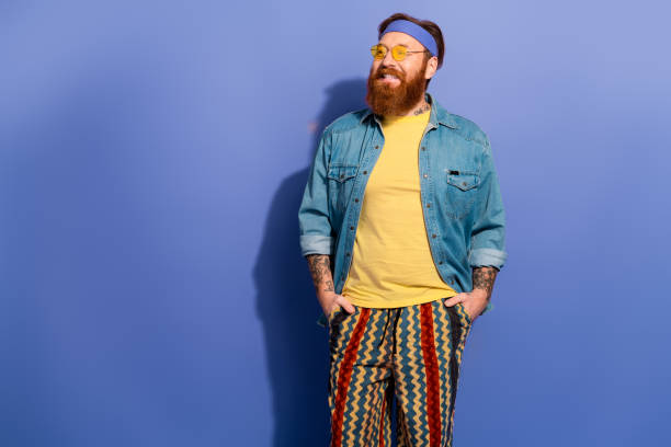 istock Confident bearded man in a yellow shirt, denim jacket, and colorful patterned pants against a violet backdrop, exuding retro hipster style 2203213229