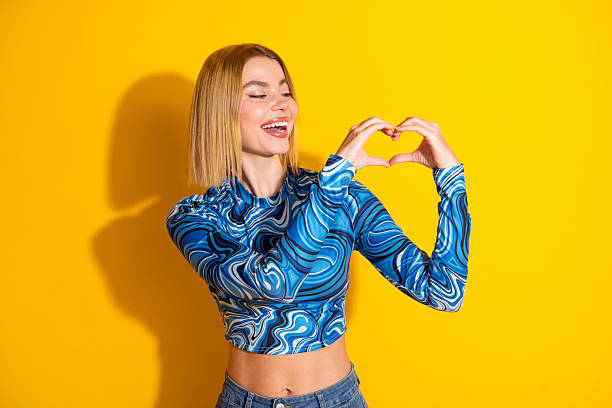 istock Smiling young woman in stylish patterned outfit making a heart shape with her hands against a bright yellow background 2203213439