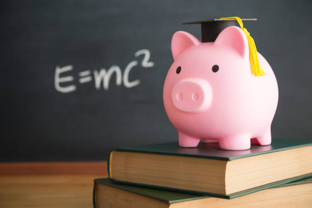 Piggy bank wears mortarboard on textbook with chalkboard in classroom.