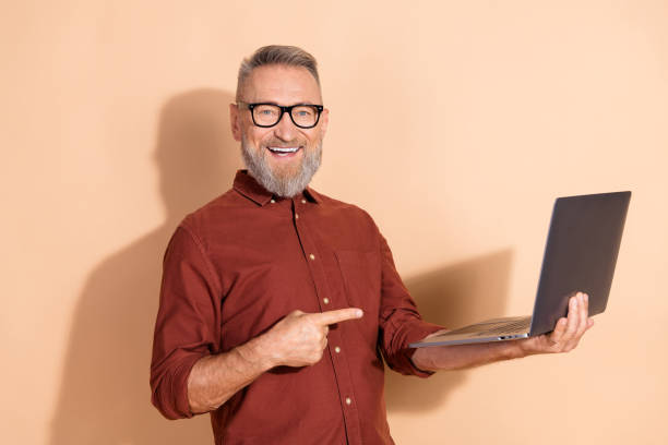 istock Smiling mature businessman holds and points at a laptop while promoting or presenting against a beige background 2203216512