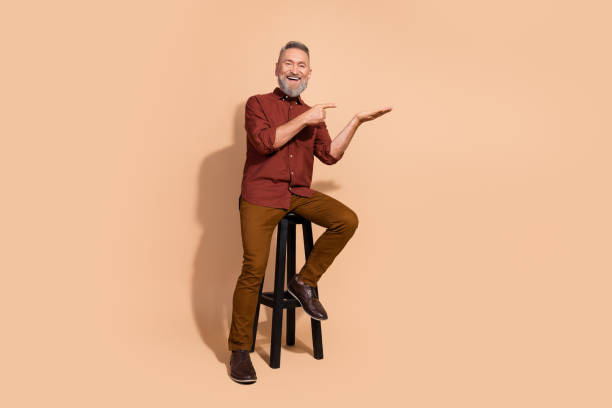 istock Elegant mature man sitting on a stool against a beige backdrop, gesturing optimistically and smiling in casual attire, ideal for concepts 2203216602