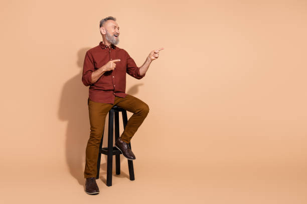 istock Handsome elderly man seated on a stool, gesturing and smiling, casual fashion in a studio against beige background 2203216603
