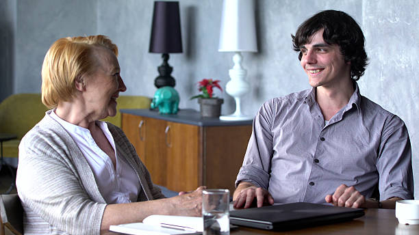 Grandson having conversation with grandmother laughing together. Portrait of smiling young man and senior woman sitting at table and talking in living room