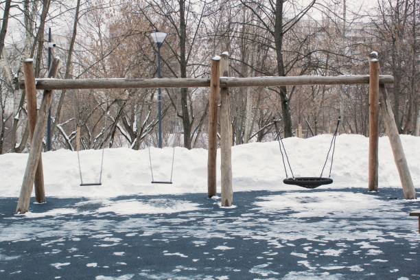 A wooden swing set stands in a snow-covered park. The set has a horizontal beam supported by four posts, with three swings hanging. The ground is a mix of snow and dark blue surfacing. Trees are visible in the background