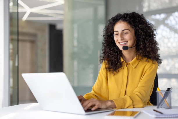 istock Joyful customer support and consulting service worker with headset smiling, using laptop for video call. Woman inside office, remotely helping customers, supporting 2203223176