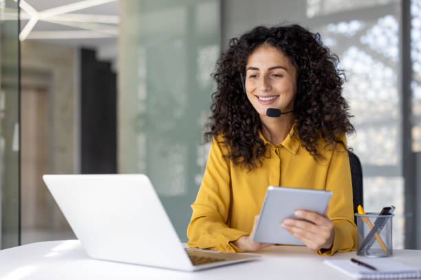 istock Woman with headset at workplace inside office holding tablet computer. Office worker of customer support and consulting service, inside office at workplace with laptop 2203223195