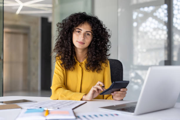 istock Confident female accountant sits at her desk with documents and laptop, using a calculator to manage company finances. 2203223238