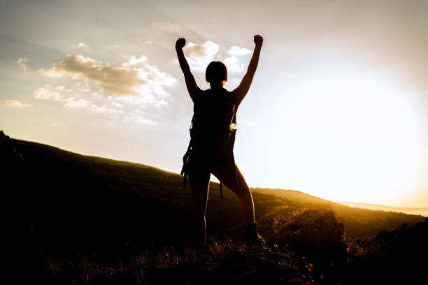 Silhouette of young hiker woman standing on a mountain top with arms outstretched