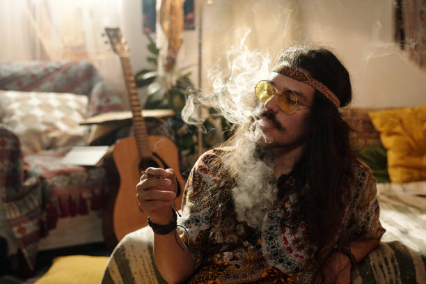 Man with long hair and sunglasses sitting in a cozy room filled with bohemian decor and musical instruments, exhaling smoke from his mouth