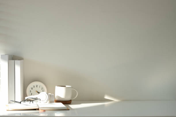 istock Minimalistic workspace featuring a white desk with books, a coffee mug, glasses, headphones, and a clock. 2203249747