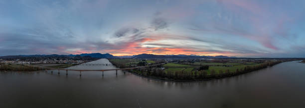 Panoramic view of Mission, British Columbia at sunset, featuring the Fraser River, lush fields, and distant mountains under a colorful sky. Captures the serene beauty of the Canadian landscape.