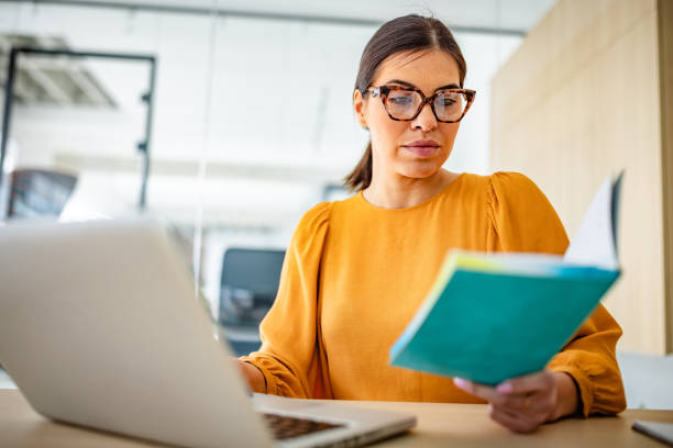istock Young woman engages in focused work at a modern office, balancing digital tasks with reading for professional growth 2203252979