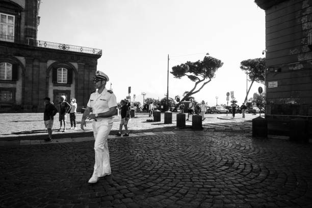 The image shows a naval officer in uniform walking on a cobblestone street in Naples, Italy. The background features historic architecture with arched windows and a group of people, likely tourists, observing the surroundings. The scene captures the authenticity of daily life in Naples, with a picturesque tree and street elements adding to the charm of the city.