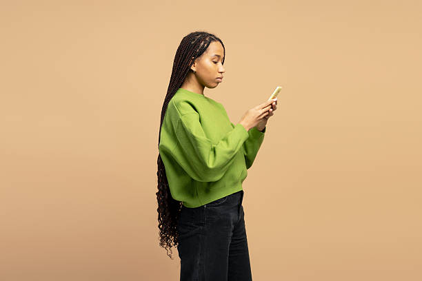 istock Young woman using mobile phone, standing in profile view on beige background 2203262282
